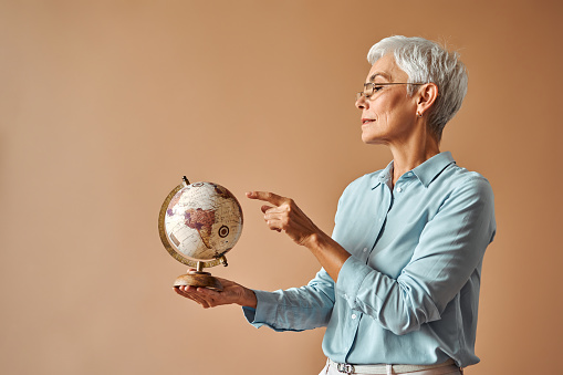 Beautiful confident short haired gray haired mature woman in blue blouse and glasses holding vintage globe choosing country for retirement travel. Rest and travel around the world, tourism.