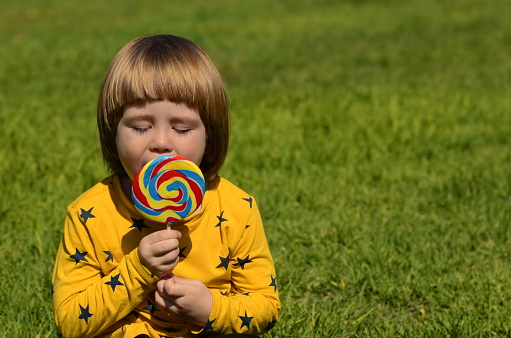 A little boy is enjoying a big candy on a stick. Multicolored lolipop. Child on a green background. Summer concept, family holidays, birthday, amusement park