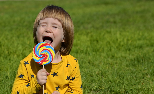 A little boy is enjoying a big candy on a stick. Multicolored lolipop. Child on a green background. Summer concept, family holidays, birthday, amusement park