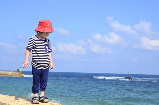 A 3 years old boy with long hair in a sailor's striped vest T-shirt with an anchor. The kid is standing on a sandy beach, in the background the sea. Concept: family vacation at sea, beach vacation