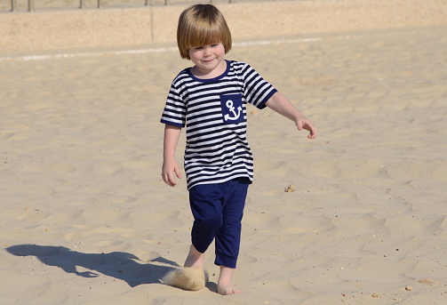 A 3 years old boy with long hair in a sailor's striped vest T-shirt with an anchor. The kid is standing on a sandy beach, in the background the sea. Concept: family vacation at sea, beach vacation