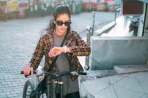 A young woman uses a bicycle to easily reach any destination within a 15-minute city of Himachal Pradesh for a sustainable and eco-friendly lifestyle. She looks at her wristwatch as she waits for someone.