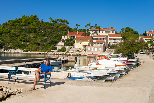 Man relaxing on the bench and looking at mobile phone in front of small boats port with Zverinac town in background.  It is small town on small island in the Zadar Archipelago as part of the Adriatic Sea coast. It is part of Croatia and Mediterranean.