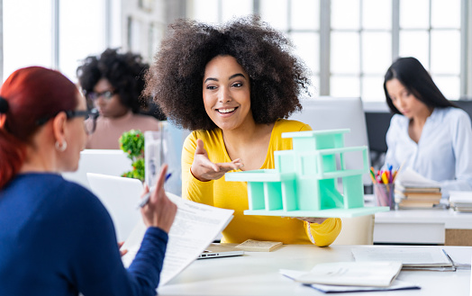 Teamwork. African american businesswoman in office, holding model of a building, having discussion with a colleague. Business Marketing Planning.