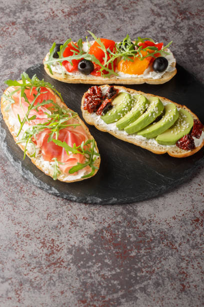 istock Three different open sandwiches with arugula, jamon, sun-dried tomatoes, avocado, cream cheese, olives and roasted peppers close-up on a slate board. Vertical 1949679717