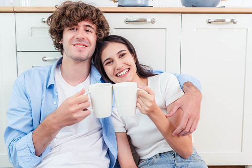 Young couple tasting coffee drinks enjoying flirt and conversation Smiling millennial European woman and man with cups of coffee sitting on floor talk in kitchen interior. Communication at home