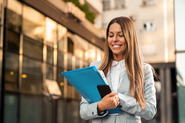 istock Successful female entrepreneur holding cellphone and document folder while walking by the office building 1949802005