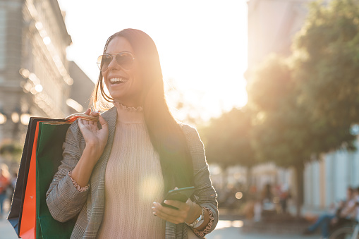 Attractive smiling woman with long hair and sunglasses holding paper shopping bags and mobile phone walking down the city street on a sunny late afternoon.