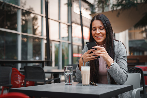 Attractive young woman sitting alone outdoors in urban environment, enjoying her drink and texting while waiting a friend to join her.