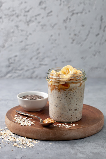 healthy diet breakfast. overnight oatmeal with chia seeds, bananas, peanut butter, honey in a glass jar on a gray concrete background