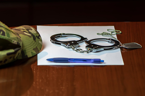 On a wooden table lay a camouflage military cap, a pair of army medallions, metal handcuffs and a blue pen with a piece of paper. Concept: law enforcement, military authority.