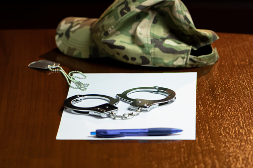 A military cap, handcuffs and army dog tags, a pen with a white sheet on a wooden table.