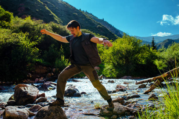 Young man hiker crosses mountain river at sunny summer day