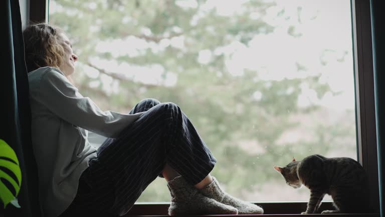 A young woman sitting on the windowsill with her cute cat