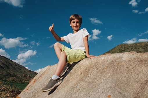 Happy boy sits on a huge stone in the mountains and gives a thumbs-up