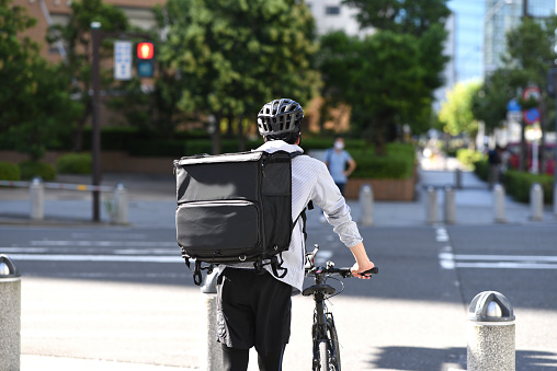 Rear view of a young Asian man holding a backpack and providing delivery service on a bicycle