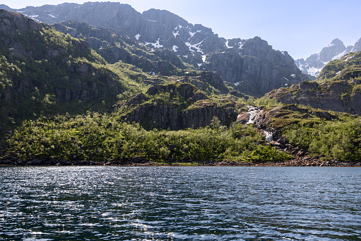 Light glints off the ripples of Trollfjorden's waters, with a delicate waterfall nestled among the craggy mountains and lush greenery of Lofoten, Norway