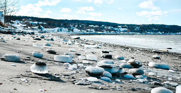The photograph captures a serene winter scene where ice-encased rocks are scattered along a snowy beach, with a blurred town visible across the water.