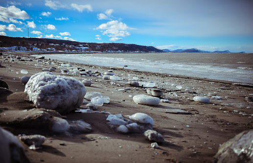 The photograph captures a serene winter scene where ice-encased rocks are scattered along a snowy beach, with a blurred town visible across the water.