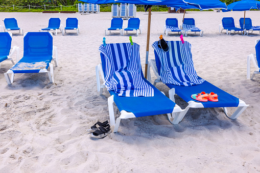 View of the sandy Miami beach with sun loungers covered with beach towels secured with clips and beach slippers left behind.
