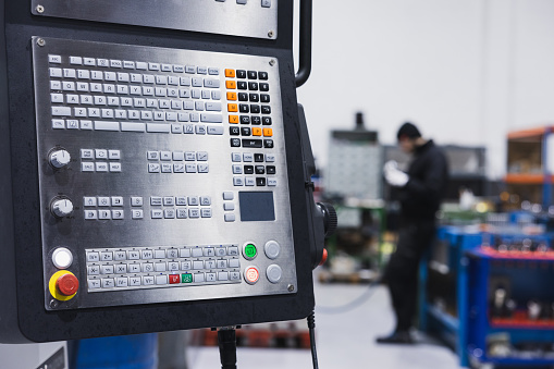 A focused view of a complex control panel of metalworking CNC milling machine, with a blurred background featuring an engineer meticulously working on machinery. Industrial concept.