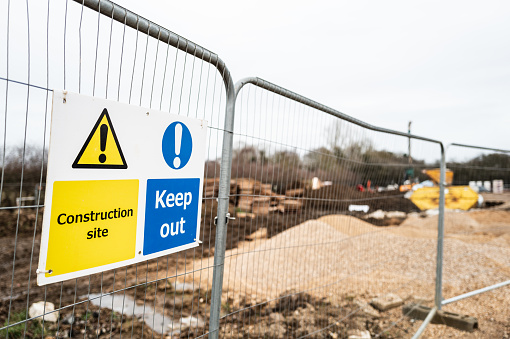 Shallow focus of a Keep Out and Construction Site sign seen on a metal fence on a new housing development in England. A skip and construction vehicles can just be seen in the distance.