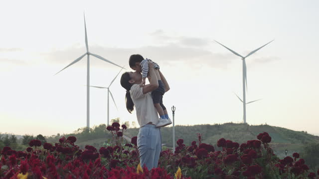 Asian mother holding her adorable son playing with pinwheel wind turbine at wind turbine energy farm exploring outdoor learning sustainable energy resource. Children outdoor learning activity experience environmental sustainability lifestyle concept.