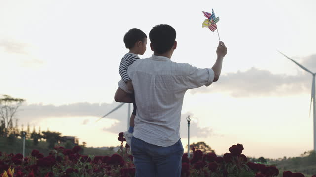 Asian father holding his adorable son playing with pinwheel wind turbine at wind turbine energy farm exploring outdoor learning sustainable energy resource. Children outdoor learning activity experience environmental sustainability lifestyle concept.