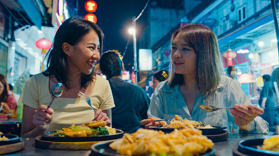 Young Asian female friends tourist enjoy eating street food on the street at night market in Bangkok, Thailand. Holiday vacation trip concept.