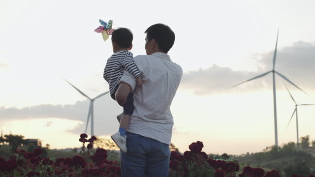 Asian father holding his adorable son playing with pinwheel wind turbine at wind turbine energy farm exploring outdoor learning sustainable energy resource. Children outdoor learning activity experience environmental sustainability lifestyle concept.