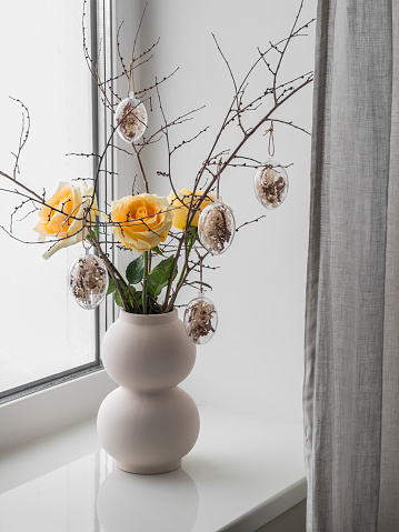 Easter bouquet of yellow roses and dry twigs with decorative glass eggs on the windowsill. Simple Easter Decor