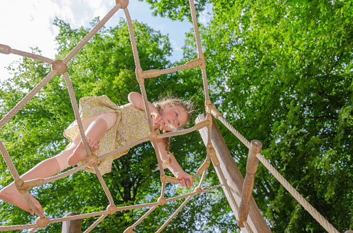 five-year-old girl on climbing frame. She is blonde with two pigtails and barefoot. Greenery and sky in background.