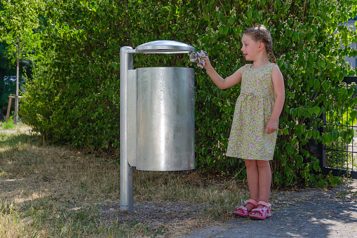 little girl throws trash into street trash can. She is blonde with long braid and wearing summer dress. As a background green bushes of blooming jasmine trees