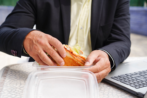 Close-up of an unrecognizable businessman eating a sandwich while working with laptop in a sidewalk cafeteria