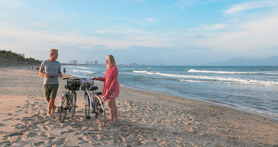 Mature couple park bicycles on beach and walk towards waves