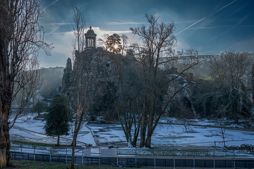 Paris, France - 01 20 2024: Park des Buttes Chaumont. View the central part of the park with footbridge, belvedere island, Temple of the Sibyl and the lake under under the snow