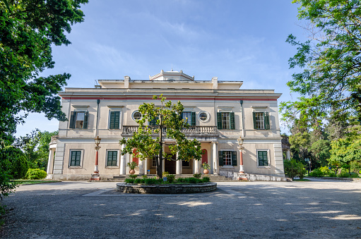Corfu, Greece - May 10 2022: View of old villa Mon Repos with columns, balcony and green wooden window shutters. Palace in a beautiful park.