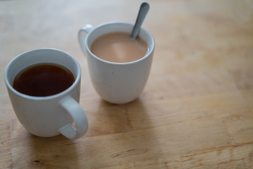 Two white mugs filled with hot tea, one with milk and a spoon, sitting on a wooden surface.