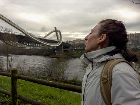 In this striking image, a solo traveler stands in awe, gazing upon the sweeping curves of a modern bridge. The overcast sky creates a dramatic backdrop, highlighting the traveler's connection with the contemporary structures and the natural surroundings. It's a moment of quiet reflection and admiration for the blend of innovation and environment.