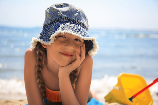 Little girl in stylish hat sunbathing on beach near sea