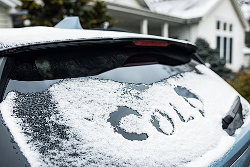 The word COLD has been sketched/scraped into the back window/windshield of a frozen, snow and ice covered generic car parked in a suburban driveway during a January cold spell. Image taken near Rochester, New York State.