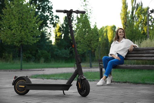 Modern electric kick scooter and woman sitting on bench in park, selective focus