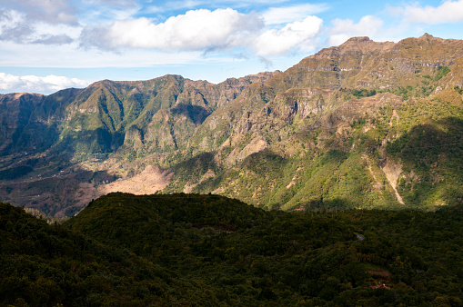 This image showcases rugged mountains, quaint villages, and meticulously carved terraces. The lush greens and earthy browns of the hills contrast with the vibrant patches of cultivated land, illustrating a harmonious blend of nature and human touch. The interplay of light adds drama, highlighting Madeira's tranquil yet dynamic beauty.