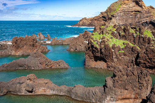 This photograph reveals the raw, unspoiled beauty of Madeira's north coast, where natural rock pools carved into the rugged shoreline. The azure waters of the Atlantic Ocean gently lap into these basaltic formations, creating a mesmerising contrast with the dark, volcanic rocks. The play of sunlight on the water's surface adds a shimmering quality, while the surrounding cliffs and greenery provide a stunning backdrop, epitomising the island's wild and natural charm.