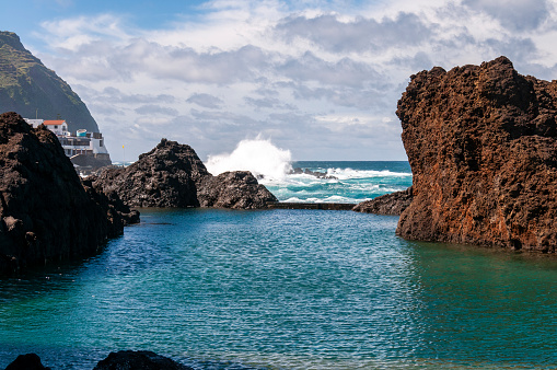 This photograph reveals the raw, unspoiled beauty of Madeira's north coast, where natural rock pools carved into the rugged shoreline. The azure waters of the Atlantic Ocean gently lap into these basaltic formations, creating a mesmerising contrast with the dark, volcanic rocks. The play of sunlight on the water's surface adds a shimmering quality, while the surrounding cliffs and greenery provide a stunning backdrop, epitomising the island's wild and natural charm.