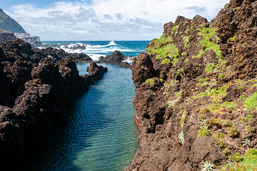 This photograph reveals the raw, unspoiled beauty of Madeira's north coast, where natural rock pools carved into the rugged shoreline. The azure waters of the Atlantic Ocean gently lap into these basaltic formations, creating a mesmerising contrast with the dark, volcanic rocks. The play of sunlight on the water's surface adds a shimmering quality, while the surrounding cliffs and greenery provide a stunning backdrop, epitomising the island's wild and natural charm.