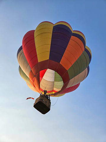 Looking up from directly below at a colorful hot air balloon as it is flying overhead with a plain blue sky as a background.