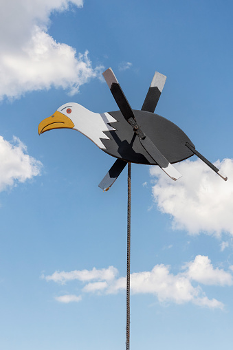 Seagull shaped wooden weathervane on a thin rod in blue sky with white clouds background. Vertical photo.