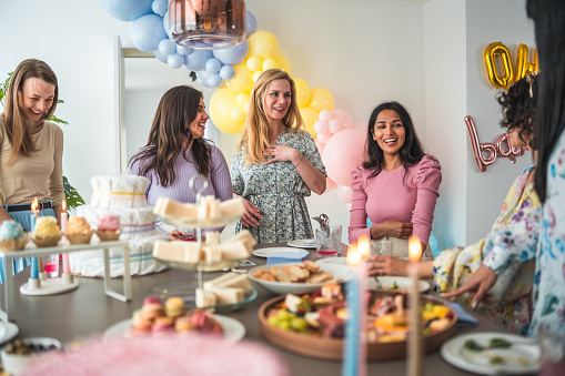 Females of diverse backgrounds gather around a baby shower table adorned with a sumptuous selection of cheeses, fruits, and baked goods.