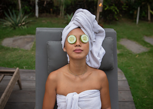 Close-up shot of Asian woman relaxing with a cucumber slice covering their eyes during facial treatment. She's sitting on a tanning bed with a wrapped towel on her head and body.